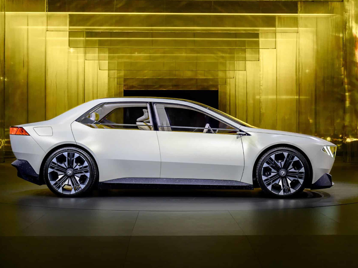 Berlina BMW de color blanco mostrada como prototipo, vista de perfil lateral, con líneas de diseño limpias y ruedas de gran tamaño, expuesta en un espacio interior iluminado con paneles dorados al fondo.