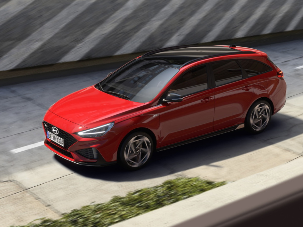 Coche compacto Hyundai i30 de color rojo circulando por una vía urbana elevada, visto desde arriba y el lateral, con carrocería alargada y techo en contraste oscuro.