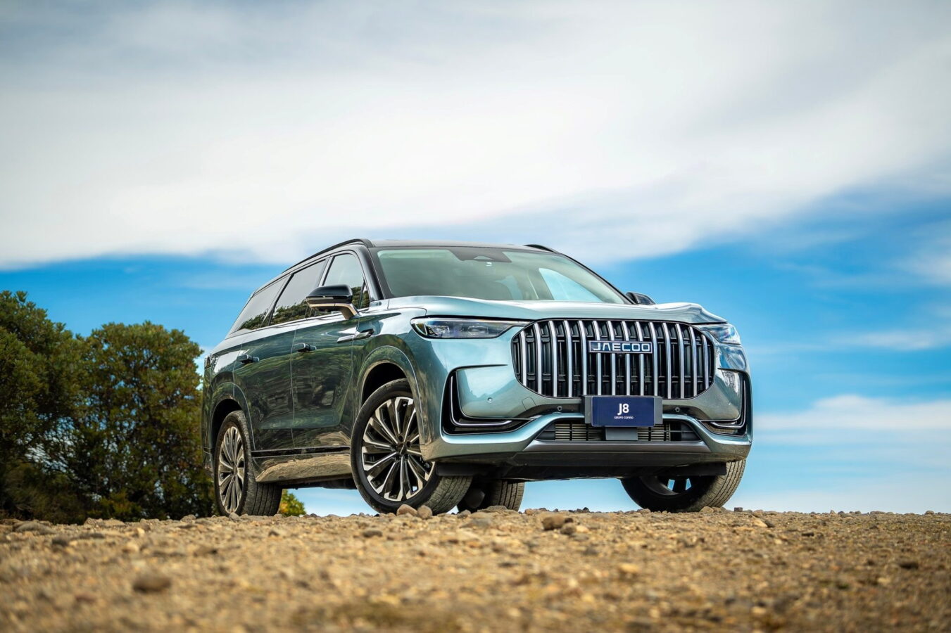 SUV Jaecoo 8 de color verde metalizado, aparcado sobre un terreno de tierra y piedras, visto desde el frontal y el lateral, con carrocería grande y parrilla vertical cromada.