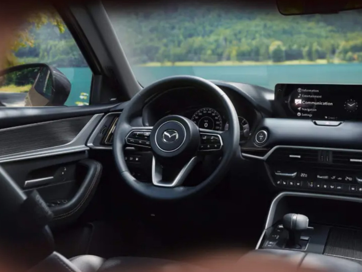 Interior del Mazda CX-60 con volante a la izquierda, salpicadero en tonos oscuros con inserciones decorativas, cuadro de instrumentos analógico y pantalla central elevada del sistema multimedia.