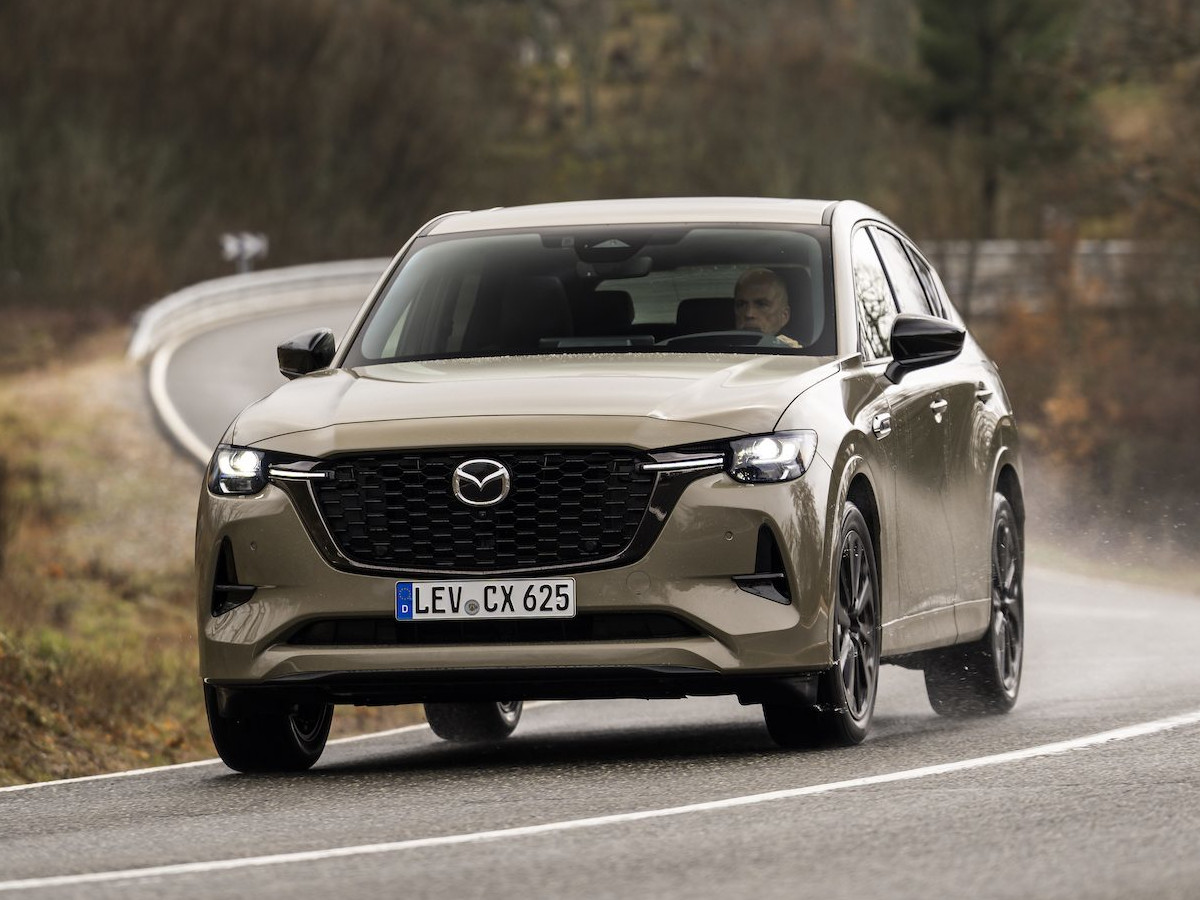 SUV Mazda CX-60 de color beige circulando por una carretera de curvas, visto desde el frontal, con el vehículo en movimiento y un entorno natural con árboles al fondo.