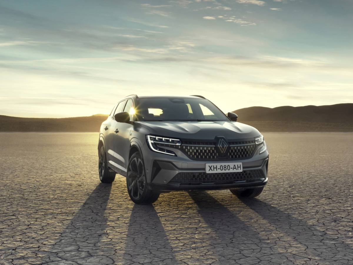SUV Renault Austral de color gris oscuro, aparcado sobre un terreno seco y abierto, visto desde el frontal, con el vehículo iluminado por la luz del atardecer y un paisaje amplio al fondo.