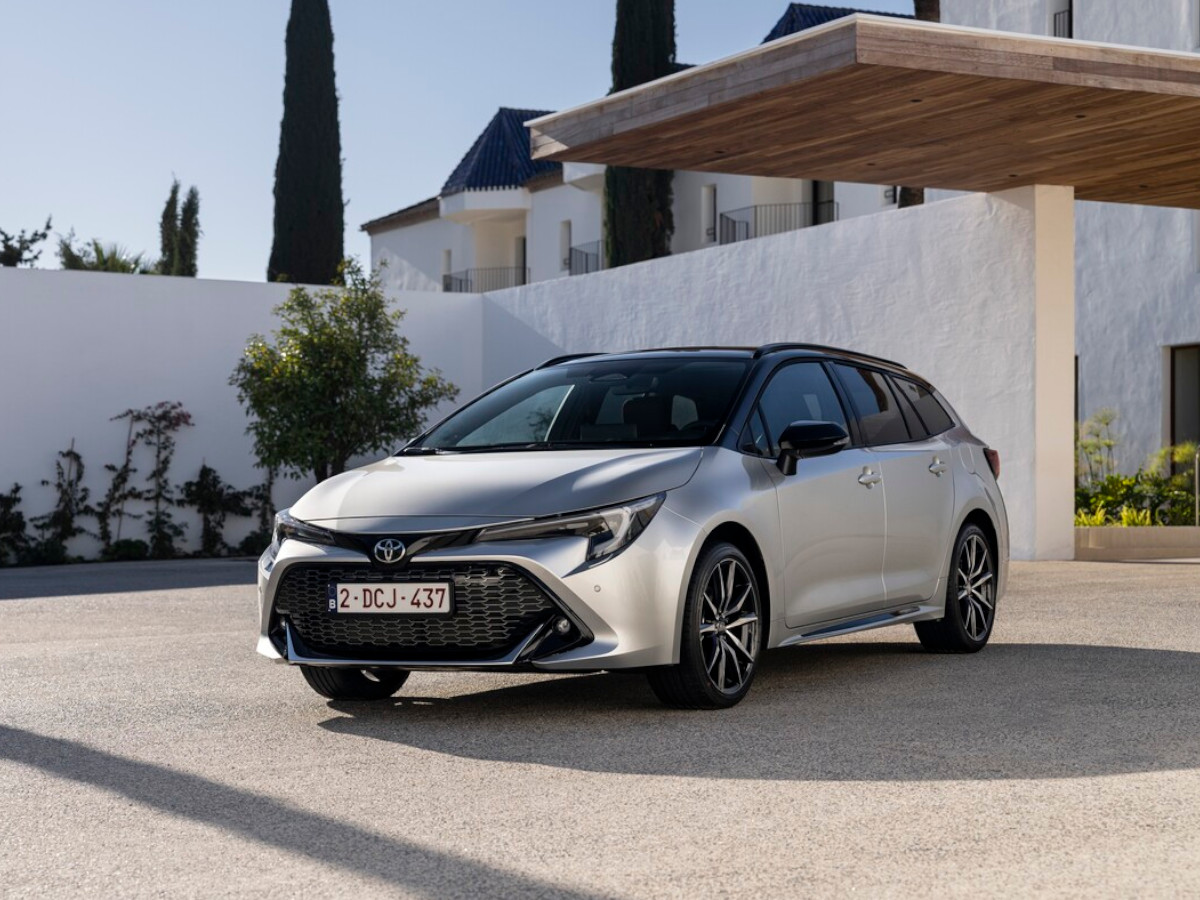 Coche familiar Toyota Corolla Touring Sports híbrido 2.0, aparcado frente a una vivienda moderna, visto desde el frontal, en color gris con techo y detalles en negro.