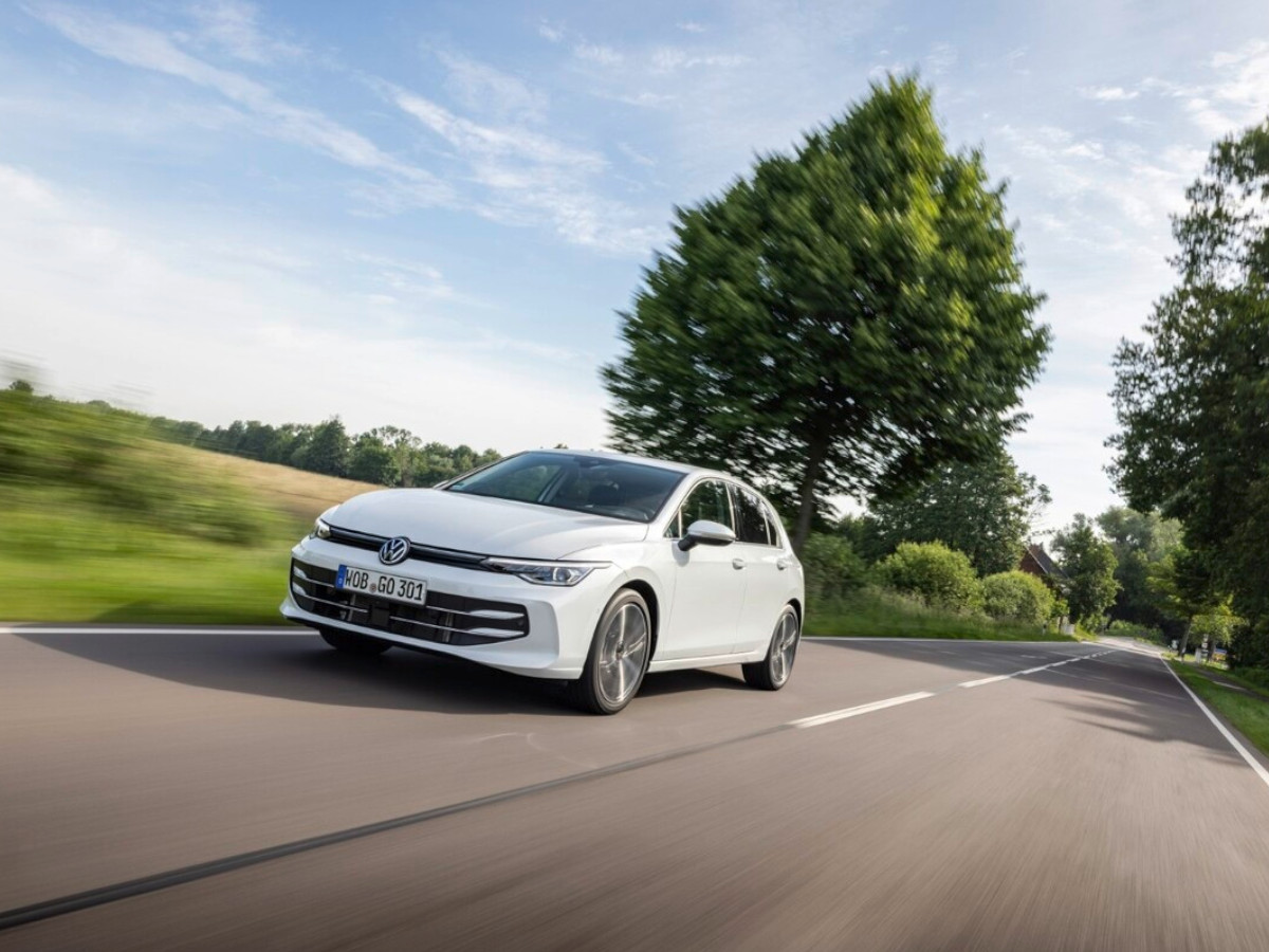 Coche compacto Volkswagen Golf de color blanco circulando por una carretera rural, visto desde el frontal en movimiento, con árboles y paisaje verde al fondo.