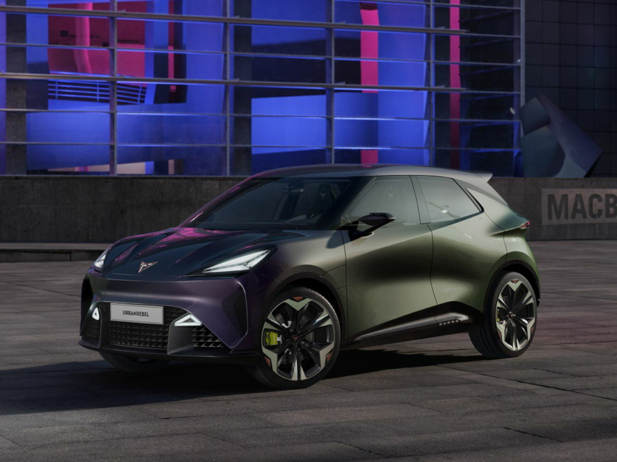 Coche eléctrico CUPRA Raval de color gris verdoso con acabado satinado, visto de frente, aparcado en un entorno urbano nocturno con iluminación LED y fachada moderna al fondo.