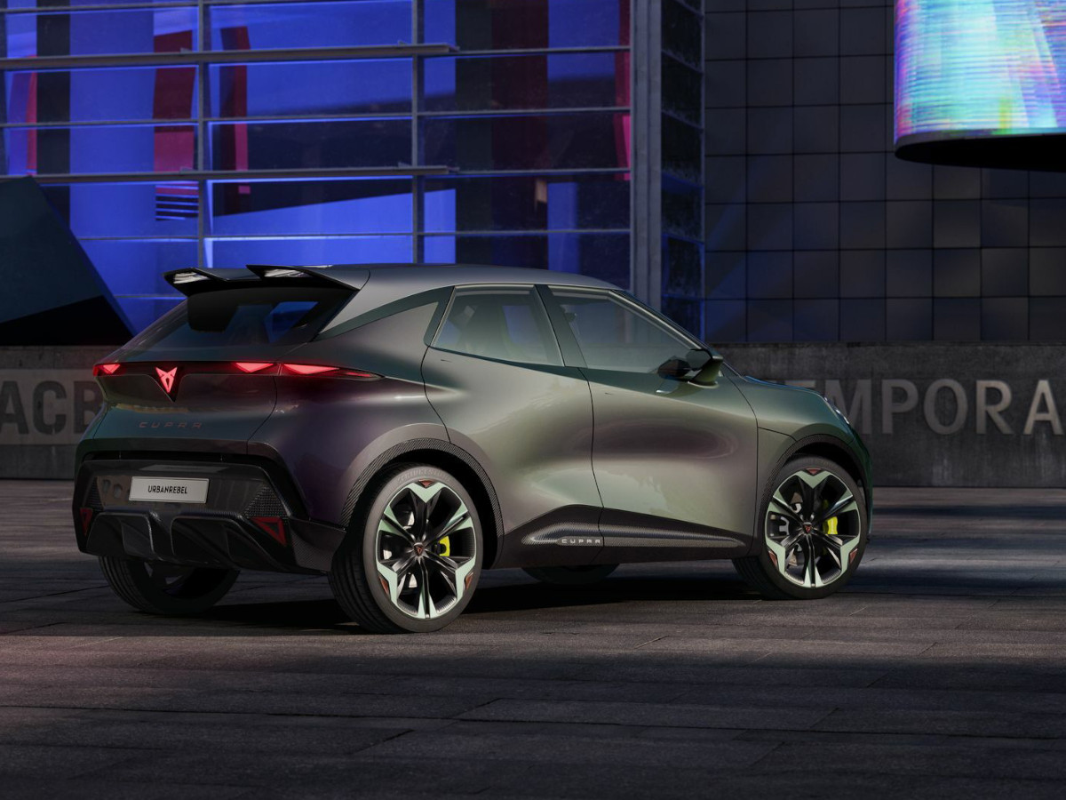 Coche eléctrico CUPRA Raval visto desde la parte trasera y lateral, con llantas deportivas, alerón trasero y firma lumínica LED, estacionado frente a un edificio moderno iluminado.