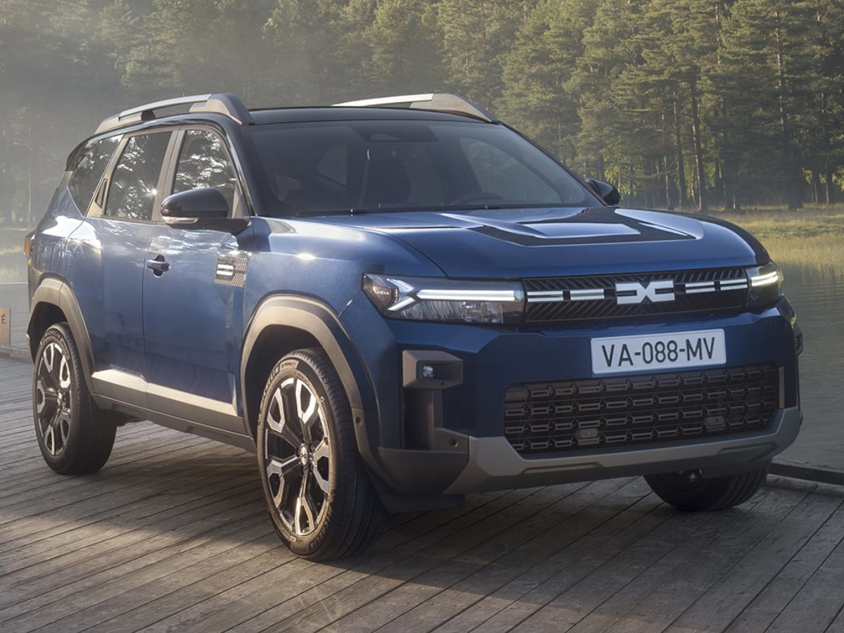 SUV Dacia Bigster de color azul oscuro visto desde el frontal y lateral, con protecciones exteriores y barras de techo, estacionado sobre una plataforma de madera junto a una zona arbolada.