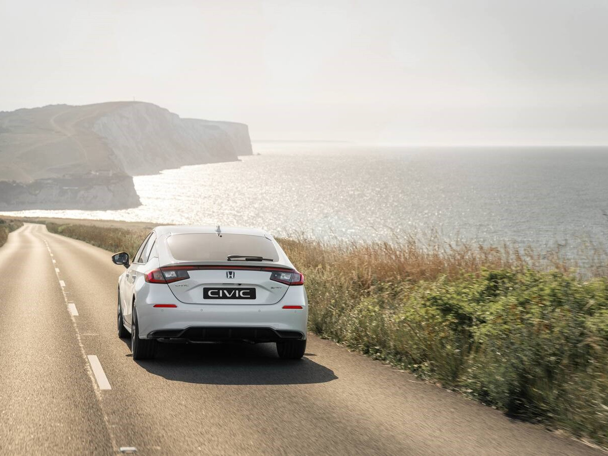 Honda Civic híbrido de color blanco visto desde la parte trasera, circulando por una carretera junto al mar, con paisaje costero y vegetación a ambos lados.