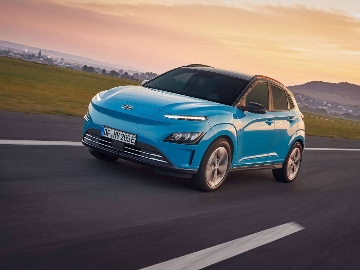 SUV eléctrico Hyundai Kona de color azul circulando por carretera abierta, visto desde el frontal y lateral, con el entorno rural y el cielo al atardecer de fondo.