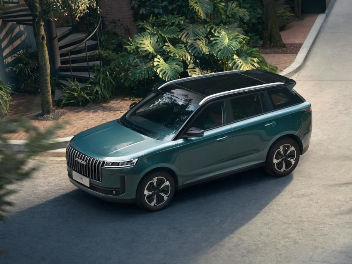 SUV Jaecoo 5 de color verde oscuro visto desde un ángulo elevado, con techo negro en contraste y llantas de diseño, circulando por una calle residencial rodeada de vegetación.