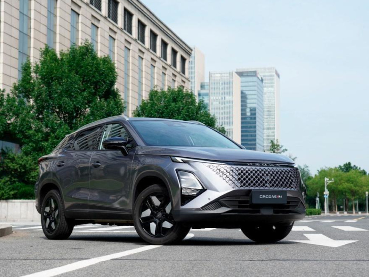 SUV OMODA 5 SHS-H de color gris oscuro visto desde el frontal y lateral, con parrilla de gran tamaño y llantas oscuras, circulando por una avenida urbana rodeada de edificios.