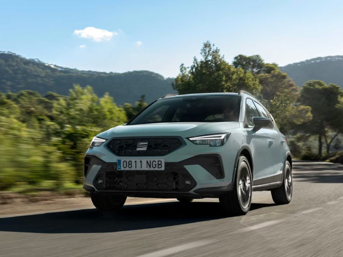 SEAT Arona de color verde claro circulando por una carretera de montaña, visto desde el frontal, con vegetación y colinas arboladas al fondo.