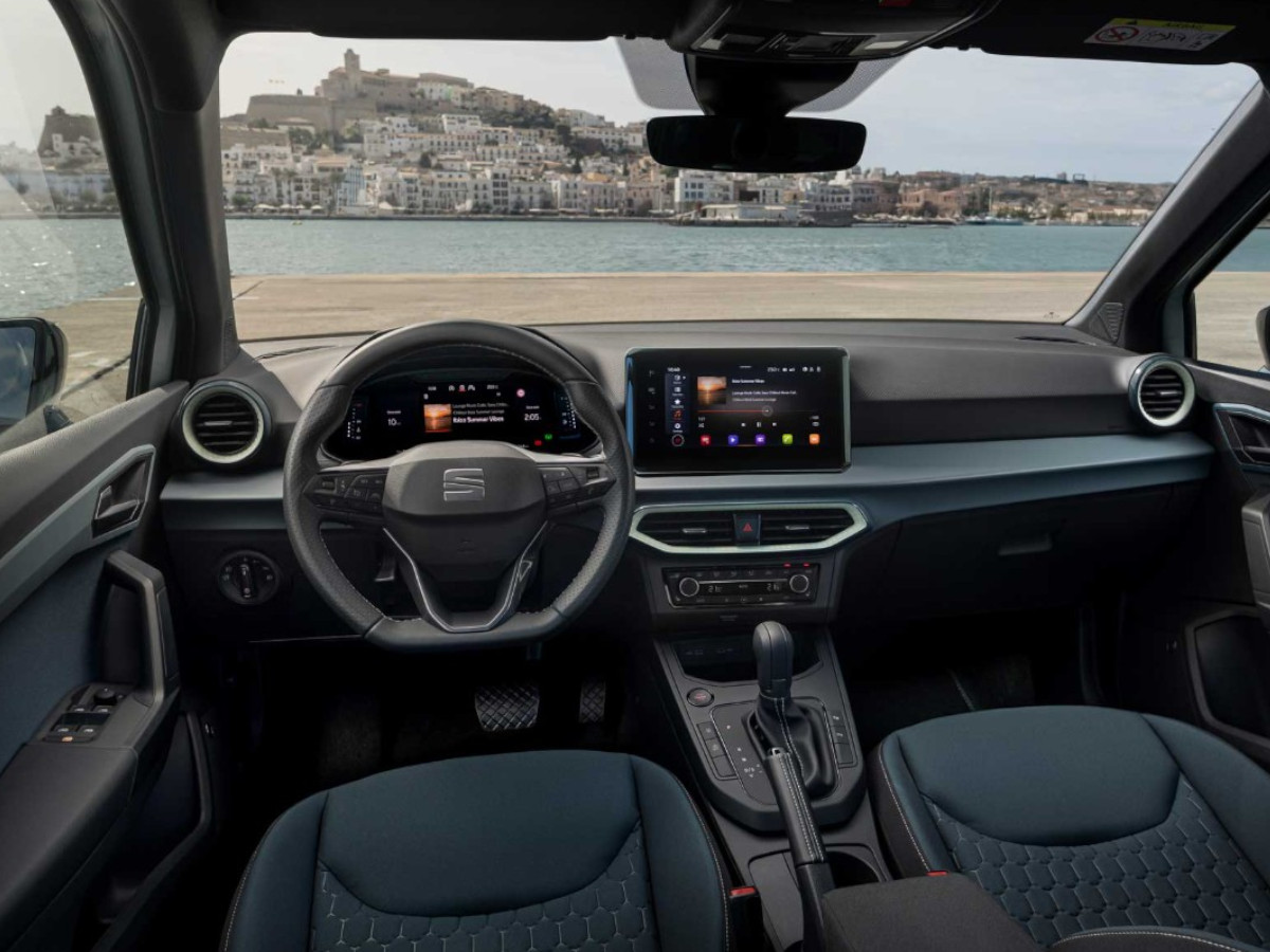Interior del SEAT Arona visto desde las plazas traseras, con volante multifunción, pantalla central flotante y salpicadero de diseño horizontal, con vistas al mar a través del parabrisas.