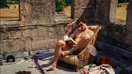 Jacob Elordi sin camiseta leyendo un libro sentado en una butaca en la película Saltburn.