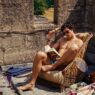 Jacob Elordi sin camiseta leyendo un libro sentado en una butaca en la película Saltburn.