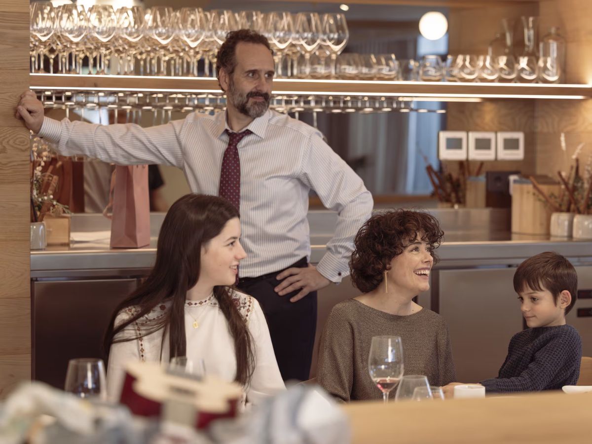 Escena de la película Los Domingos con una comida familiar. El padre vestido arreglado con camisa y corbata, la hija y la madre sentadas sonriendo.