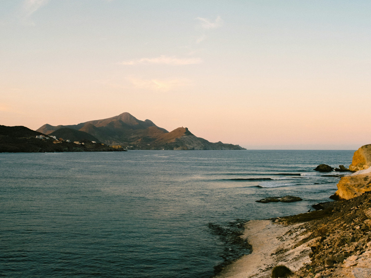 Litoral de la costa de Almería con el mar en calma, pequeñas olas llegando a la orilla, colinas áridas al fondo y luz suave de atardecer sobre el agua.