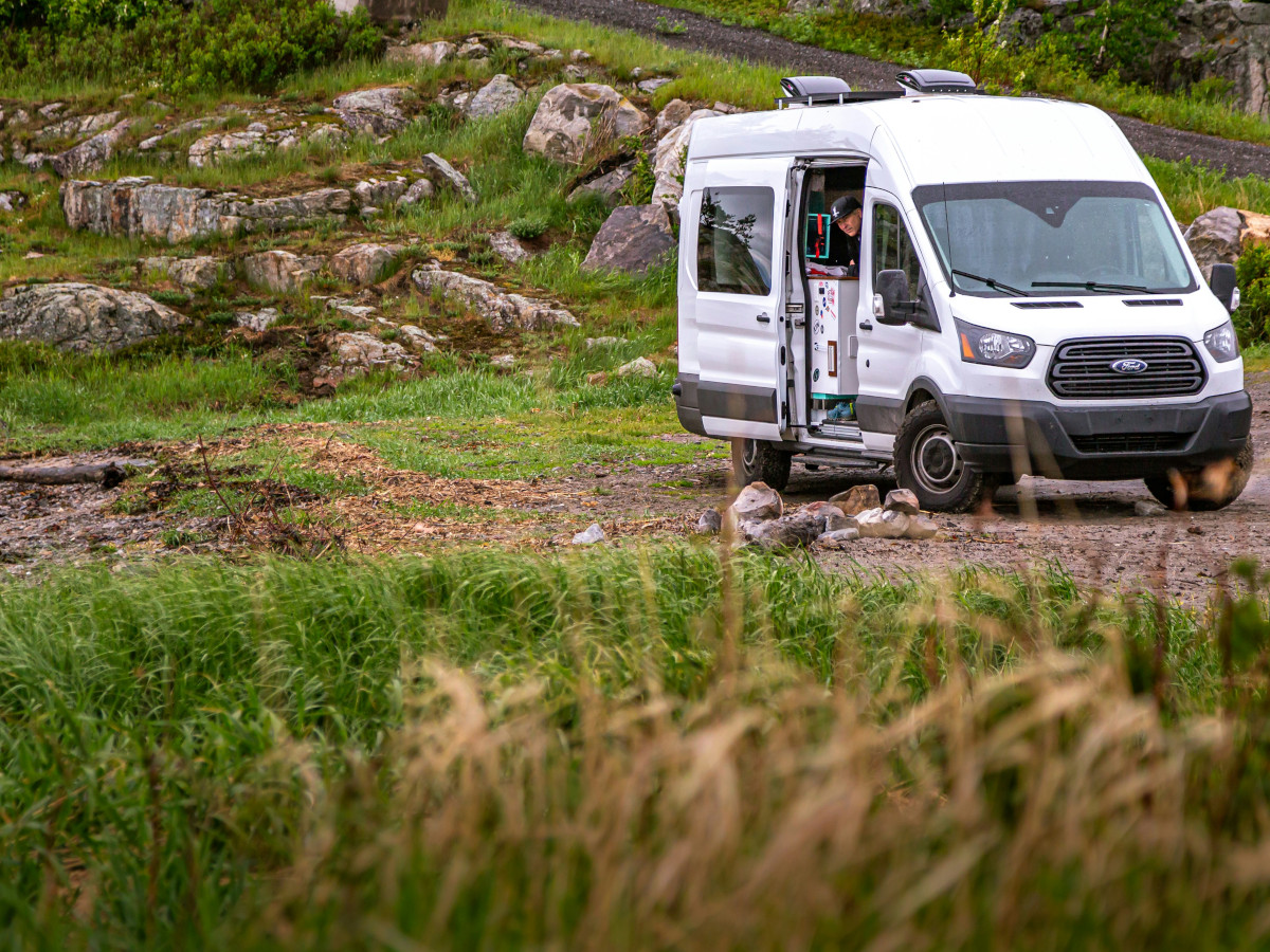 Furgoneta camper blanca estacionada en un camino de tierra rodeado de vegetación y rocas, con la puerta lateral abierta y una persona en el interior, en un entorno natural de montaña.