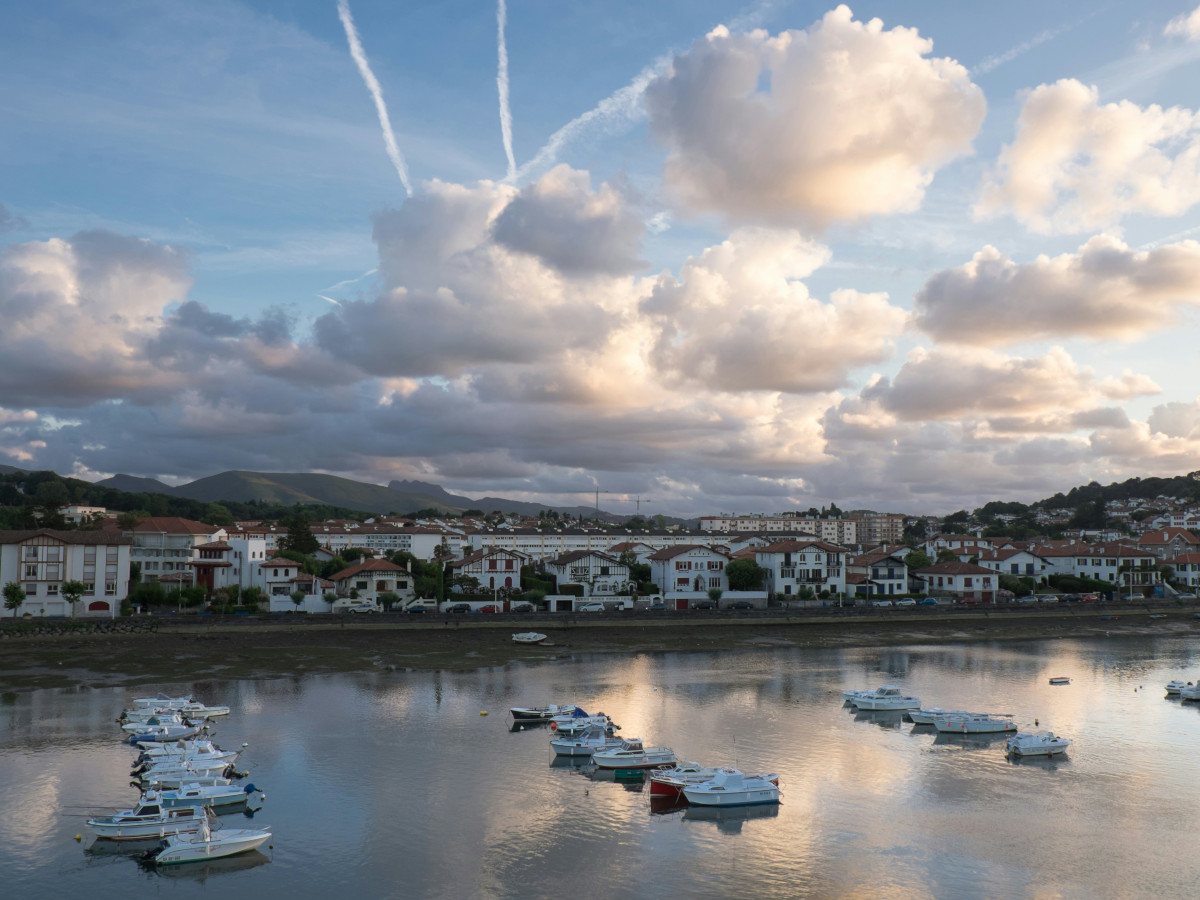 Vista de un pueblo costero de Galicia con casas blancas y tejados oscuros junto a una ría, varios barcos pequeños amarrados en el agua y colinas verdes al fondo bajo un cielo parcialmente nublado.