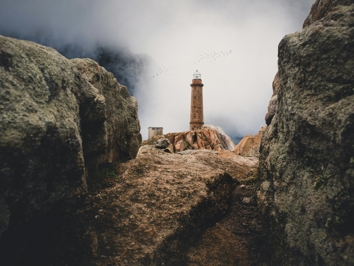 Faro de piedra situado sobre un promontorio rocoso en la costa gallega, rodeado de grandes rocas y con niebla baja y cielo nublado al fondo, típico paisaje atlántico de la Costa da Morte.