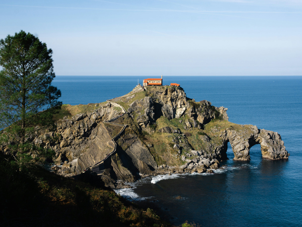 Ermita de San Juan de Gaztelugatxe situada sobre un islote rocoso frente al mar Cantábrico, unida a la costa por un puente de piedra y un camino serpenteante, con el océano azul de fondo.