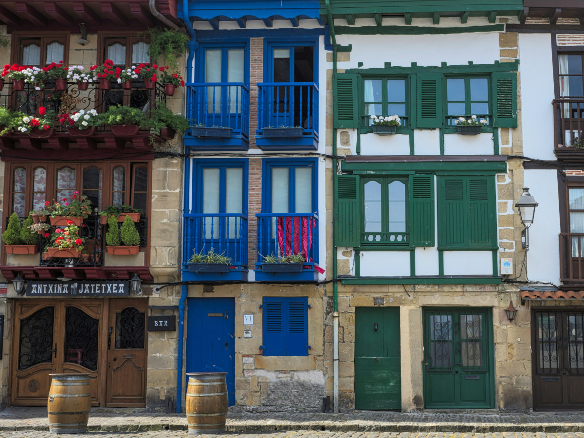 Fachadas de casas tradicionales del País Vasco con balcones de madera pintados en colores azul, verde y marrón, macetas con flores y puertas de acceso a bares y viviendas en una calle empedrada.