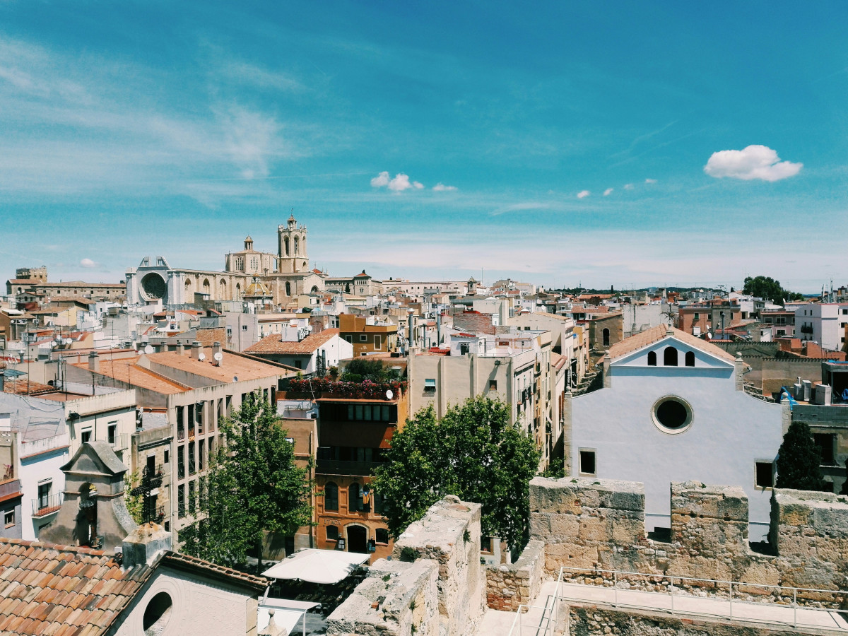 Vista panorámica del casco histórico de Tarragona con edificios de tonos claros y tejados de teja, murallas de piedra en primer plano y la catedral destacando sobre el conjunto urbano bajo un cielo despejado.