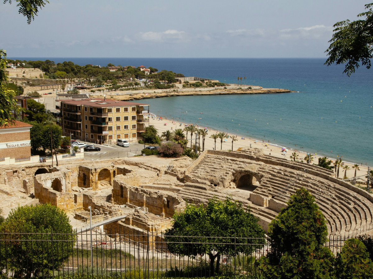 Anfiteatro romano de Tarragona visto desde lo alto, con gradas de piedra junto a la playa y el mar Mediterráneo al fondo, edificios costeros y zona urbana alrededor.
