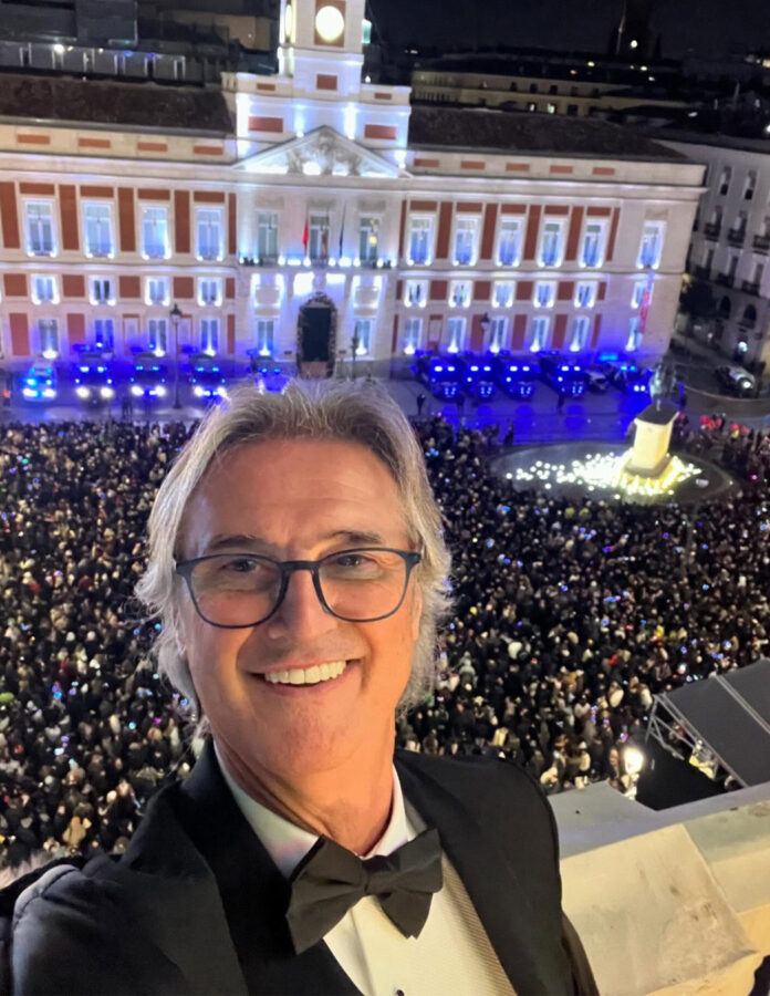 Selfie de Poty Castillo con esmoquin negro frente a una Puerta del Sol abarrotada en Nochevieja.