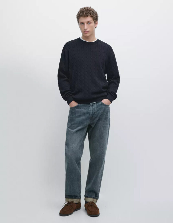 Modelo vestido con un jersey de punto trenzado azul marino, vaqueros y nauticos de ante color camel.