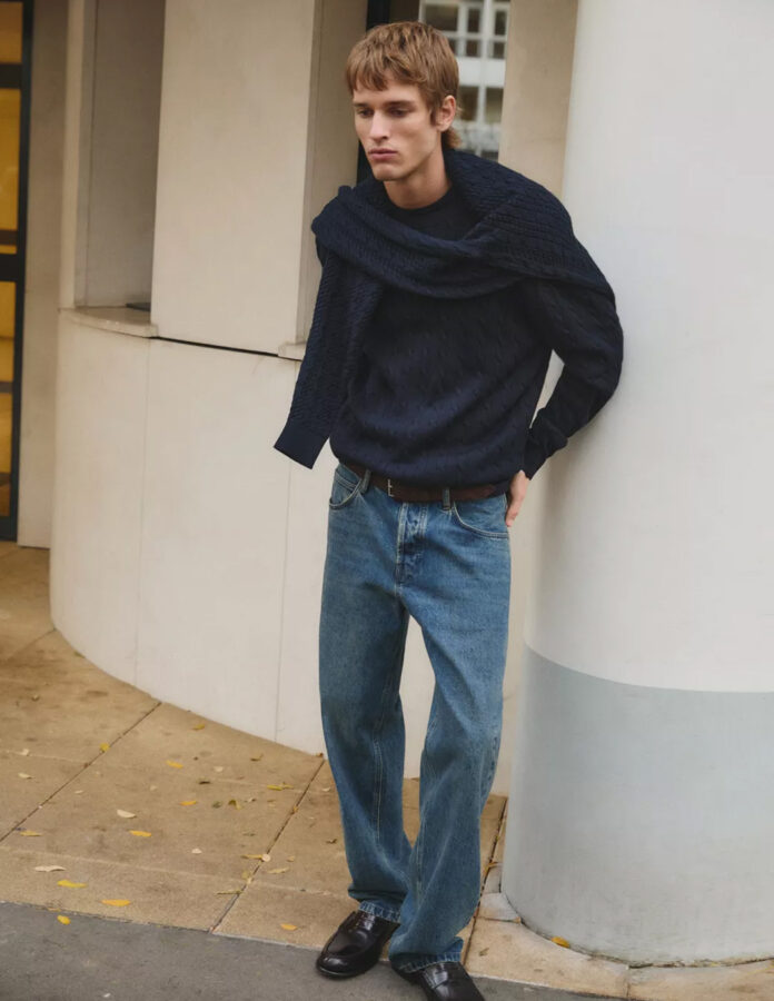 Modelo Jeray Allgayer vestido con un jersey de punto azul marino y otro jersey sobre los hombros, unos vaqueros y unos mocasines negros.