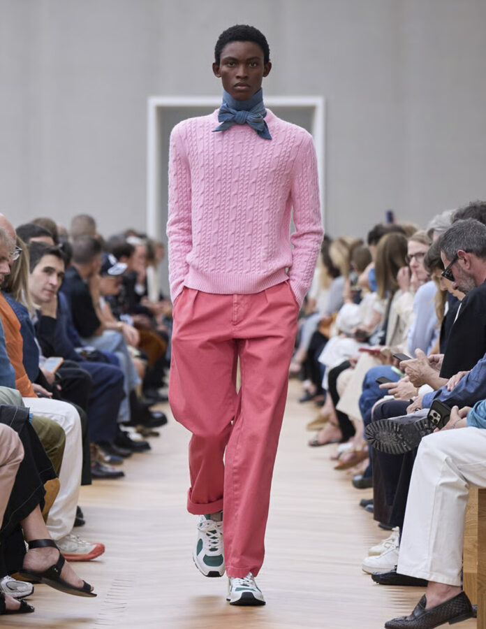 Modelo masculino en pasarela vistiendo prendas rosas superpuestas, con chaqueta amplia, pantalón a juego y detalles en capas, dentro de un desfile de moda primavera-verano.