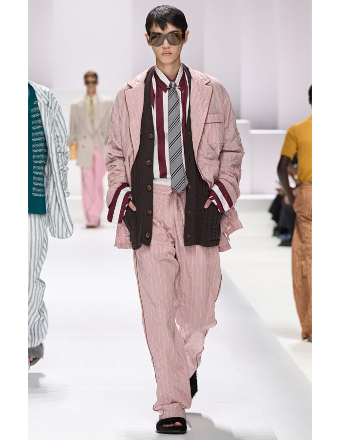 Modelo masculino desfilando con traje rosa claro de chaqueta y pantalón, camisa a rayas burdeos y blanca, corbata gris y gafas de sol, sobre una pasarela blanca.