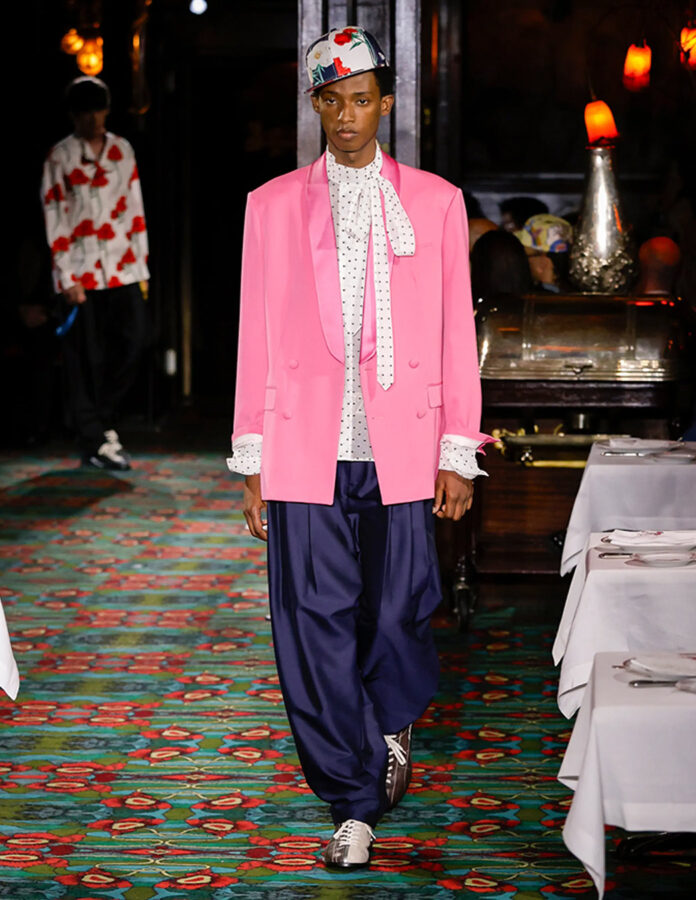 Modelo masculino caminando por una pasarela interior con americana rosa, camisa blanca con lunares, lazada al cuello, pantalón azul marino ancho y gorra estampada.