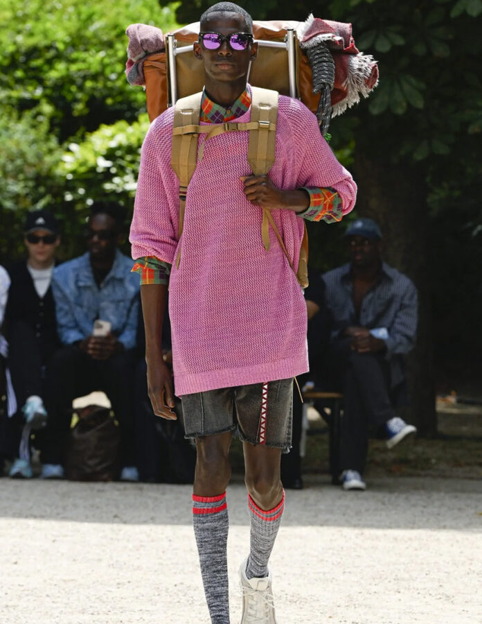 Modelo masculino al aire libre con jersey rosa de punto largo, camisa de cuadros visible en cuello y mangas, pantalón corto vaquero, calcetines altos, zapatillas y mochila grande a la espalda.