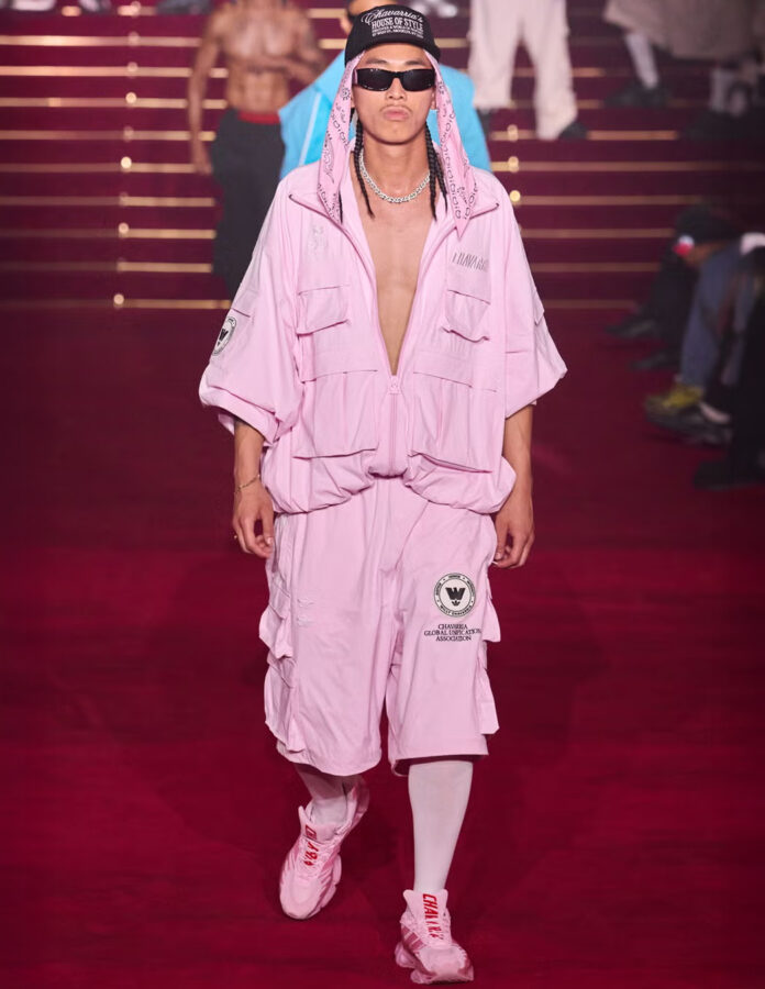 Modelo masculino desfilando con conjunto rosa completo de chaqueta y pantalón de tejido ligero, combinado con prendas superpuestas y calzado oscuro, en una pasarela de moda.