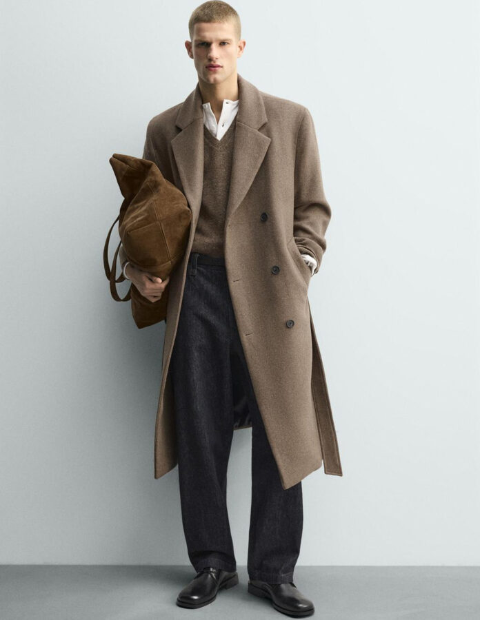 Modelo masculino viste un abrigo largo cruzado de color camel con cinturón, combinado con jersey marrón, camiseta blanca de cuello panadero, vaqueros oscuros rectos y mocasines negros.