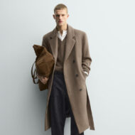 Zara tiene el abrigo camel con cinturón que vas a ver por todas partes este invierno 2026 Hombre de pie con abrigo largo camel cruzado con cinturón, llevado abierto, con look informal de invierno formado por jersey de lana, vaqueros rectos y zapatos negros.