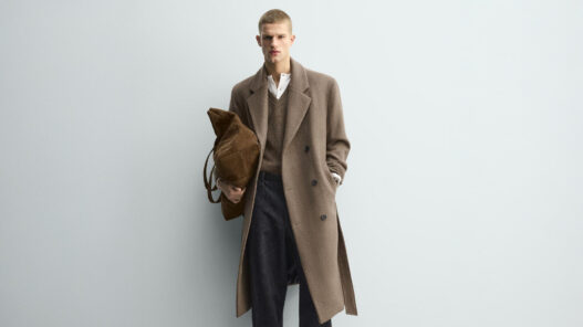 Hombre de pie con abrigo largo camel cruzado con cinturón, llevado abierto, con look informal de invierno formado por jersey de lana, vaqueros rectos y zapatos negros.