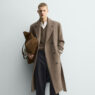 Hombre de pie con abrigo largo camel cruzado con cinturón, llevado abierto, con look informal de invierno formado por jersey de lana, vaqueros rectos y zapatos negros.