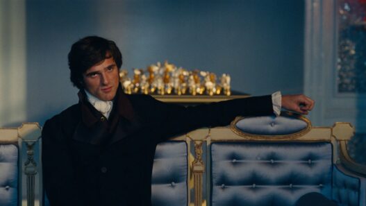 Jacob Elordi sentado en un sofá con cara seria y caracterizado como Heathcliff en Cumbres borrascosas.