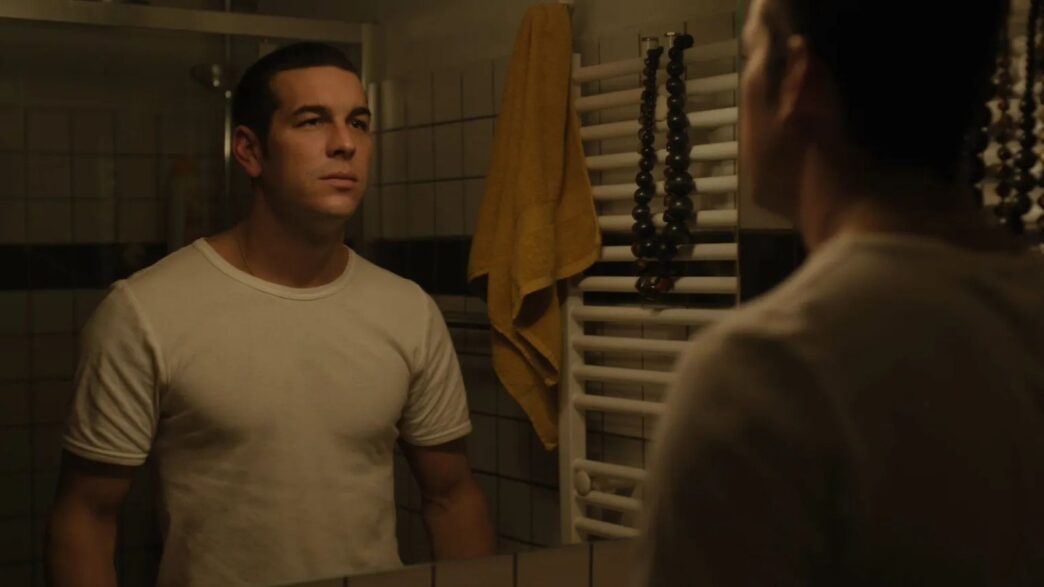 Mario Casas mirándose al espejo con una camiseta de manga corta blanca en una escena de la película Muy Lejos.