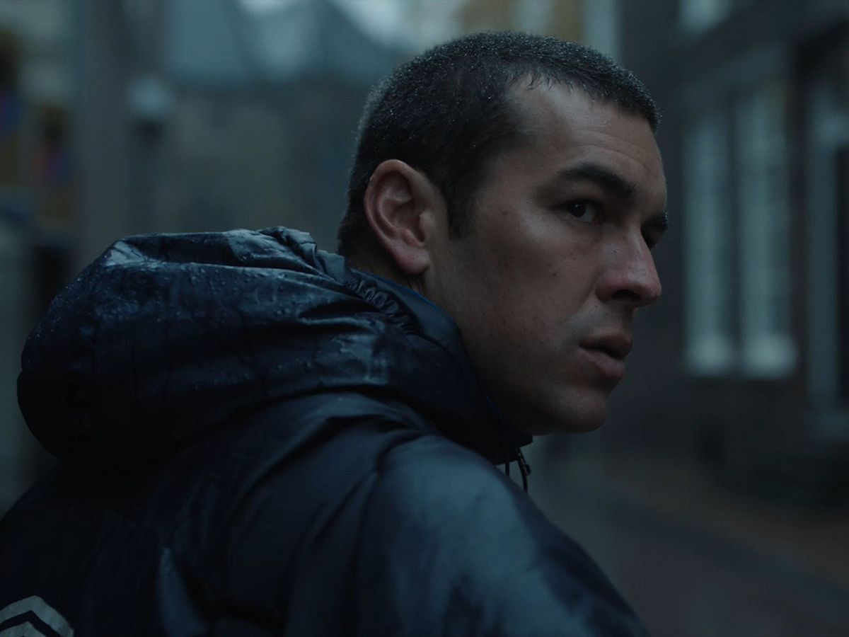 Mario Casas con el pelo rapado y un chubasquero azul en una escena de lluvia en la película Muy Lejos.