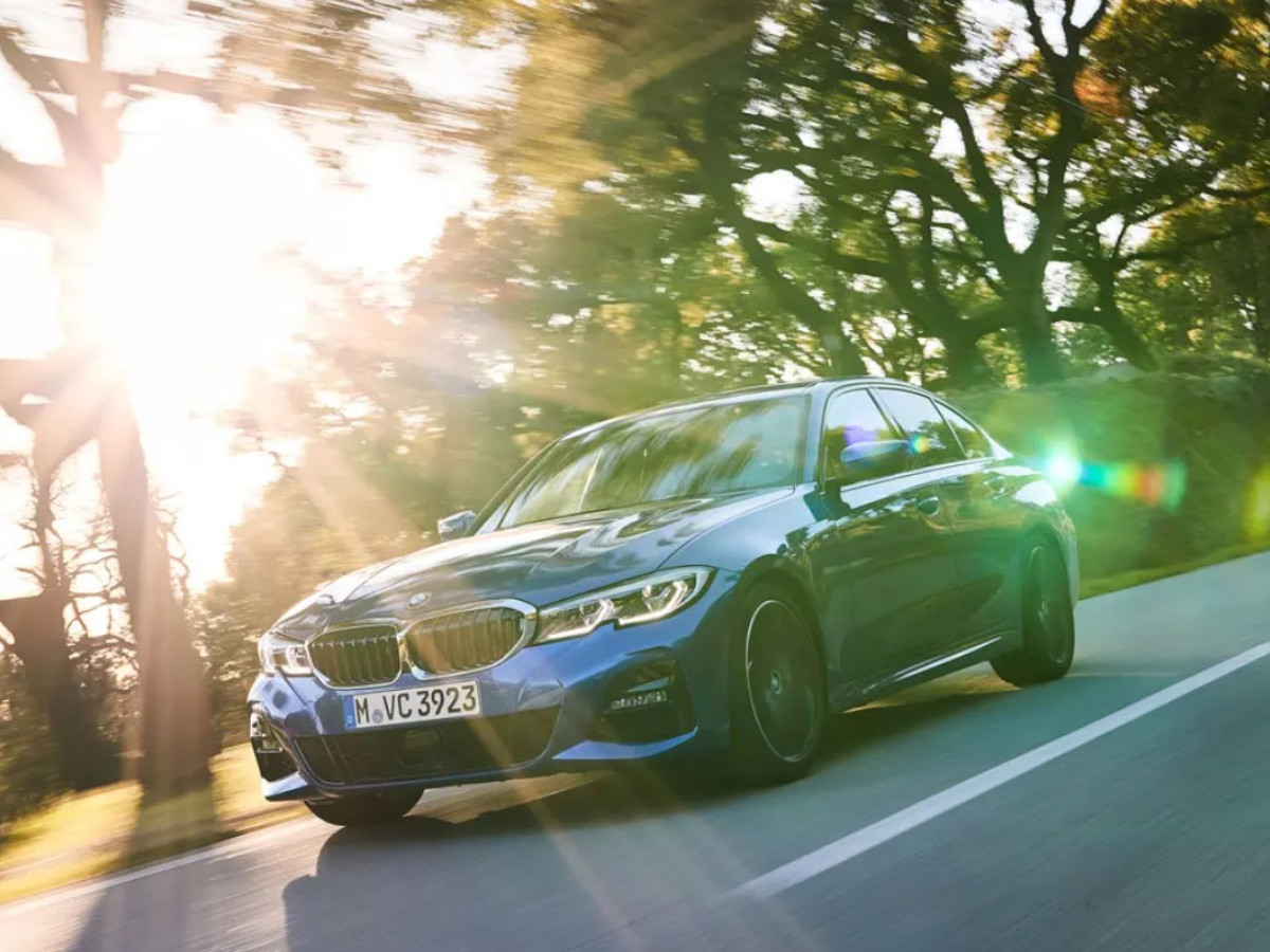 BMW Serie 3 de color azul circulando por una carretera asfaltada rodeada de árboles, visto desde el frontal izquierdo con luz de atardecer.