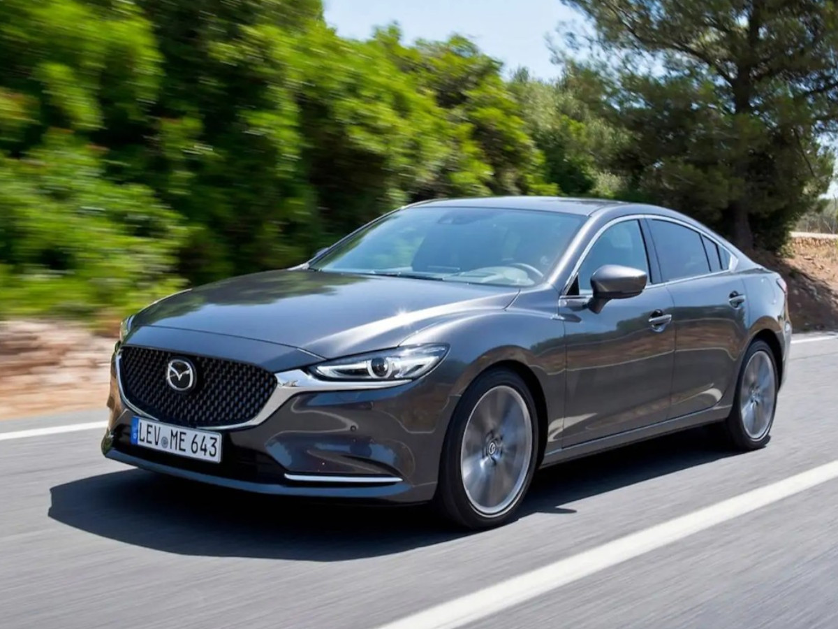 Mazda 6 de color gris oscuro circulando por una carretera asfaltada entre árboles, visto desde el frontal izquierdo en movimiento.