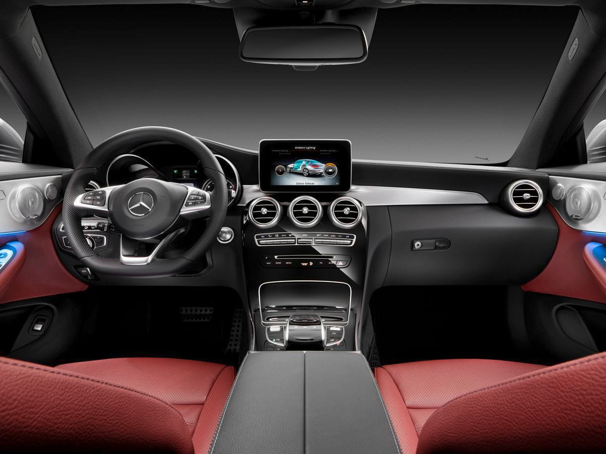 Interior de un Mercedes-Benz Clase C visto desde los asientos traseros, con volante multifunción, salpicadero negro con molduras metálicas, pantalla central y asientos tapizados en cuero rojo.