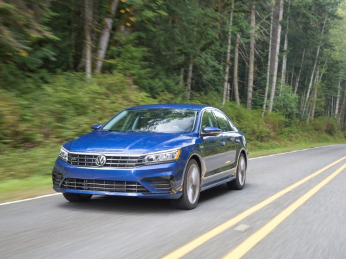 Volkswagen Passat azul circulando por una carretera secundaria rodeada de bosque, visto desde el frontal izquierdo en movimiento.