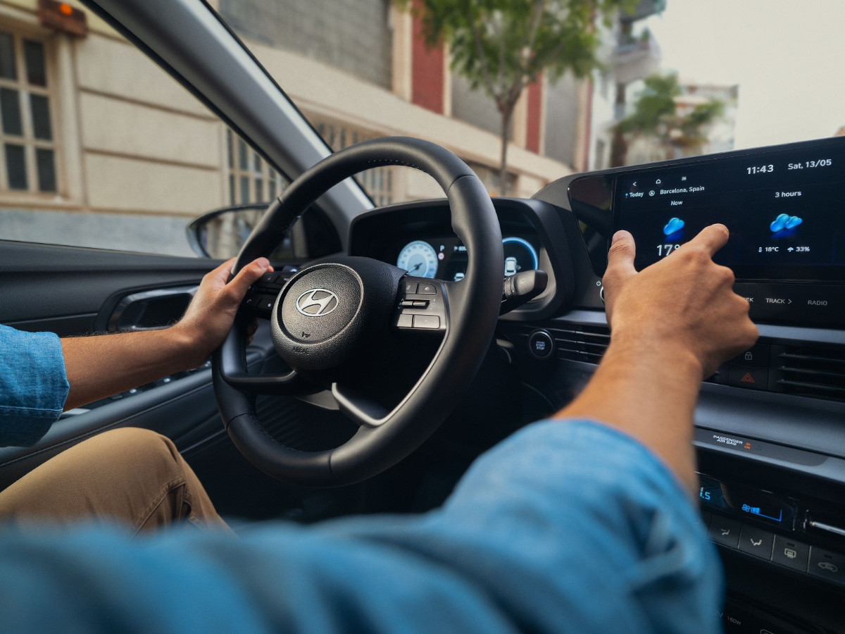 Vista desde el asiento del conductor del interior de un Hyundai i20, con manos sobre el volante y pantalla central táctil encendida mientras circula por una calle urbana.