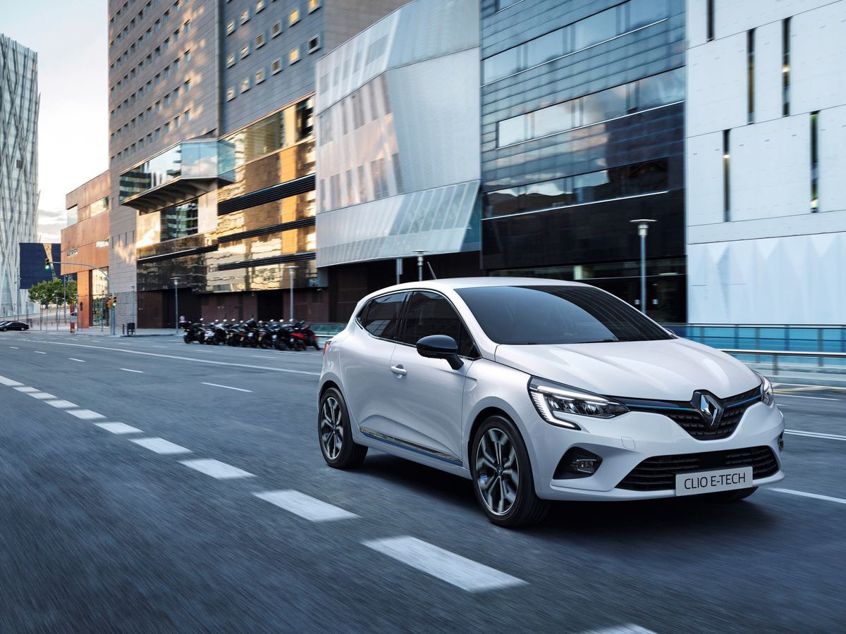 Renault Clio E-Tech de color blanco circulando por una avenida urbana moderna, visto en tres cuartos delanteros, con edificios contemporáneos al fondo.