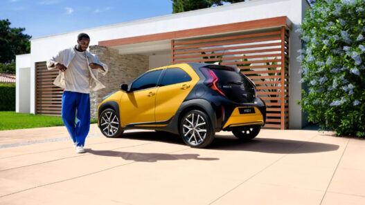 Toyota Aygo X Cross de color amarillo y negro aparcado en el exterior de una vivienda moderna, con una persona caminando junto al coche en un entorno residencial.